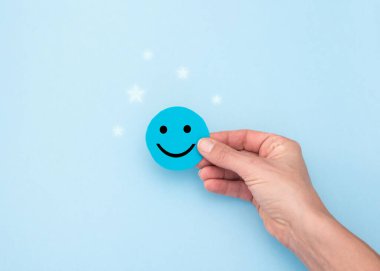 Müşteri memnuniyeti ve olumlu geri bildirim kavramı. Mavi arka planda parıltılı mavi bir memnuniyet simgesini tutan bir el.