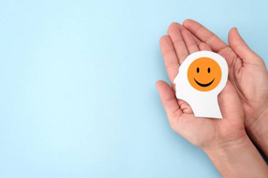 Mavi arka planda gülümseyen turuncu emojili kağıt insan kafasını tutan ellere yakın çekim