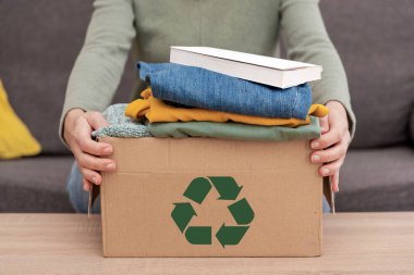 Kanepeye oturmuş, elinde geri dönüşüm kutusuyla masada bağış yapmak için kitap ve giysiler olan birinin fotoğrafını çek.