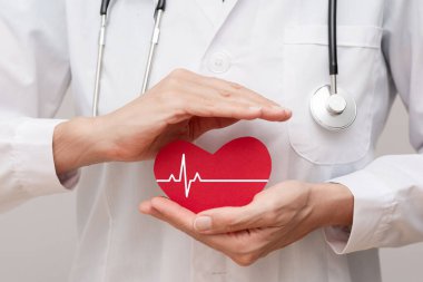 Beyaz önlüklü ve steteskoplu bir doktorun elinde elektrokardiyogram hattı, kardiyoloji sağlık sigortası ve sigorta konsepti olan kırmızı bir kalp var.