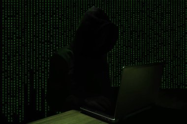 Kapüşonlu Hacker yeşil kodlu bir dizüstü bilgisayar kullanıyor.