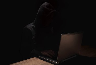 Kapüşonlu Hacker 'ın silueti koyu arkaplanda dizüstü bilgisayar kullanıyor.