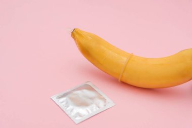 Prezervatifin kapağını muza, ambalajını da pembe arka plana, güvenli seks ve doğum kontrol kavramına yakın tut.