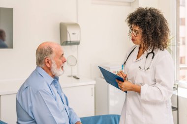 Kadın Afro doktor hastane odasında kıdemli hastaları dinlerken not alıyor.