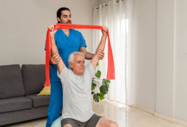 Erkek Fizyoterapist, evde ve pilates topunda direnç bandıyla egzersiz yapan yaşlı bir adama yardım ediyor.