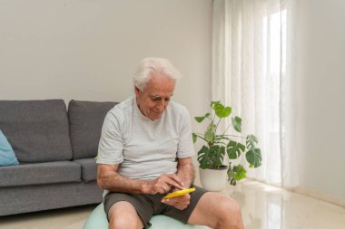 Spor salonunda otururken akıllı telefon kullanan yaşlı bir adam, emeklilikte fizyoterapi ve sağlık için evde çalışmanın tadını çıkarıyor.