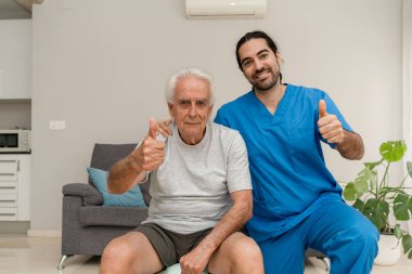 Mavi üniformalı erkek fizyoterapist ve kameraya gülümseyen ev egzersizleri sırasında baş parmak hareketi yapan yaşlı hasta.