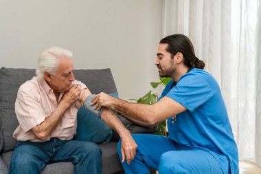 Mavi üniformalı erkek hemşire evdeki kanepede oturan yaşlı adamın kan basıncını ölçüyor.