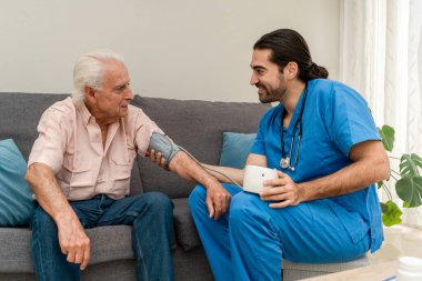 Evde mavi üniformalı erkek hemşire, yaşlı adamın kanepede oturup kan basıncını ölçüyor.