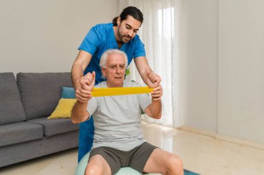 Erkek Fizyoterapist, evde sarı direniş bandıyla egzersiz yapan yaşlı bir adama yardım ediyor.