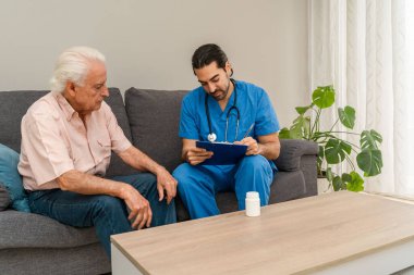 Mavi üniformalı ve steteskoplu bir erkek hemşire evde yaşlı bir adama yardım ediyor.