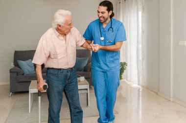 Mavi üniformalı gülümseyen erkek fizyoterapist evde rehabilitasyon sırasında koltuk değnekli yaşlı adama yardım ediyor.