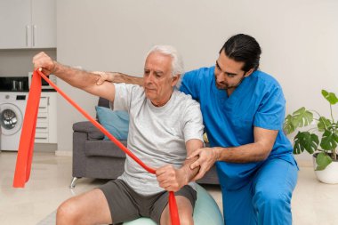 Evde direnç bandıyla egzersiz yapan kıdemli bir adam, rehabilitasyon ve hareket kabiliyetinin geliştirilmesi için fizyoterapistin yardımıyla