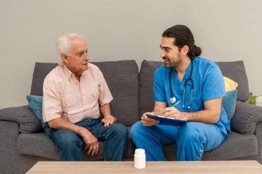 Erkek doktor, panoya not tutarken yaşlı bir adamı evinde ziyaret ediyor.