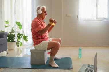 Son sınıf öğrencisi evde egzersiz yapıyor. Rezistans bandı ve kulaklıkla dizüstü bilgisayar kullanıyor.