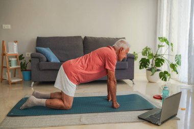 Bilgisayardaki online eğitim dersinin ardından evde yoga egzersizleri yapan yaşlı bir adam.