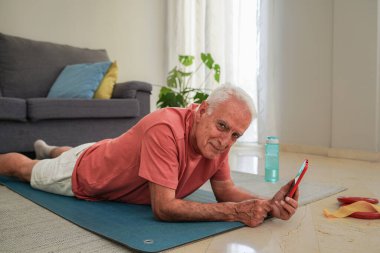 Yaşlı bir adam yoga minderinde dinlenirken akıllı telefon kullanıyor ve kameraya gülümsüyor.