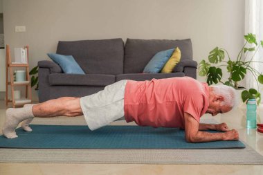 Yaşlı bir adam oturma odasında yoga minderinin üzerinde kalas egzersizi yapıyor.