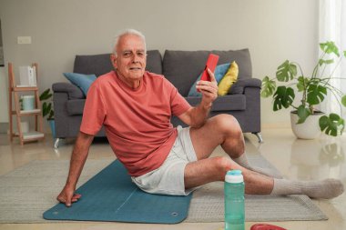 Yaşlı adam akıllı telefon kullanıyor. Evde egzersiz paspası üzerinde oturuyor. Kameraya gülümsüyor.