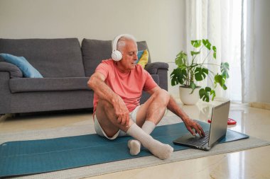 Evde egzersiz yaparken kulaklıklı yaşlı bir adam dizüstü bilgisayar kullanıyor.