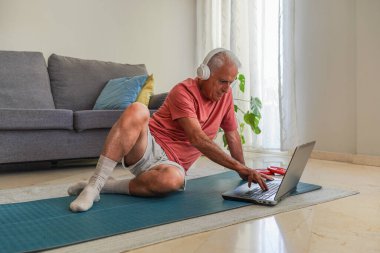 Emekli adam yoga minderinde oturmuş kulaklık takıyor evde online fitness eğitimi için dizüstü bilgisayar kullanıyor.