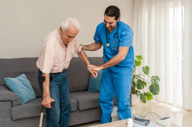 Mavi üniformalı fizyoterapist rehabilitasyon sırasında yaşlı bir adamın koltuk değneği kullanmasına yardım ediyor.