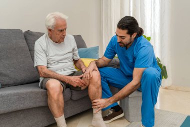 Mavi üniformalı bir erkek fizyoterapist, yaşlı adamın ağrılı dizini evde muayene ediyor. Bakım ve rehabilitasyon.
