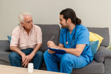 Mavi üniformalı erkek doktor ev ziyareti sırasında panoya not alan yaşlı bir adamı ziyaret ediyor.