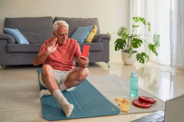 Yoga minderinde oturan yaşlı bir adam evde online antreman için akıllı telefonu kullanırken birini selamlıyor.