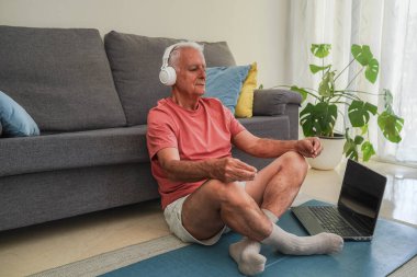 Üst düzey bir adam evde kulaklıkla bir minder üzerinde meditasyon yapıyor, akıl sağlığını geliştiriyor.
