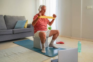 Evde elastik bantla egzersiz yapan yaşlı bir adam online fitness dersini laptopta takip ediyor.