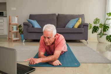 Yaşlı bir adam yoga minderinde ev çalışması için dizüstü bilgisayar kullanıyor.