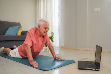 Oturma odasında yoga yapan yaşlı bir adam dizüstü bilgisayarda beden eğitimi dersini kullanıyor.