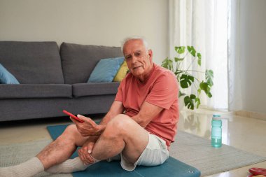 Evde akıllı telefon tutarak egzersiz yapan yaşlı bir adam yoga minderinde dinleniyor.