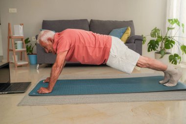 Evde dizüstü bilgisayardaki beden eğitimi dersini takiben yoga minderinde tahta egzersizi yapan yaşlı adamın yan görüntüsü.