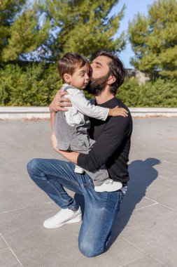 Genç baba, oğlunu parkta kollarında tutarken alnından öpüyor.