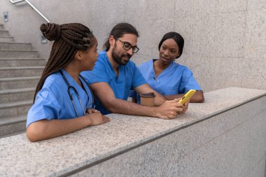 Çok etnik çeşitlilikteki tıp öğrencileri hastane binasının önünde akıllı telefon kullanıyorlar.