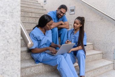 Üç farklı tıp öğrencisi dizüstü bilgisayar kullanıyor, kahve molası veriyor ve üniversite merdivenlerinde oturuyor.