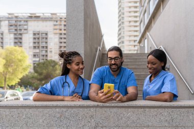 Mavi üniformalı üç tane çok ırklı tıp öğrencisi hastanenin önünde akıllı telefon kullanıyor.