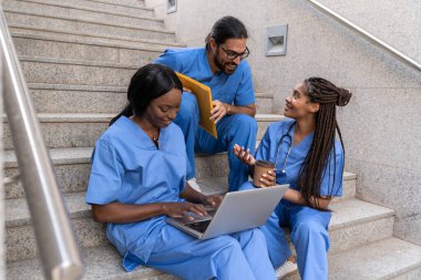 Mavi üniformalı çok ırklı tıp öğrencileri dizüstü bilgisayarlarını kullanarak ve üniversite basamaklarında fikirlerini paylaşarak birlikte çalışıyorlar.
