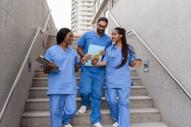 Mavi önlüklü çok ırklı tıp öğrencileri merdivenlerden iniyor, akıllı telefon kullanıyor ve konuşuyorlar.