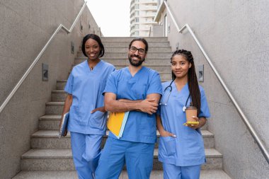 Bir grup çok ırklı mavi önlüklü tıp öğrencisi üniversite merdivenlerinde kameraya gülümsüyor.