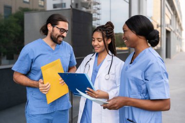 Bir grup çok ırklı doktor ve üniformalı hemşireler hastanenin dışındaki hasta kayıtlarını inceliyor.