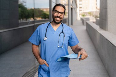 Mavi önlüklü ve steteskoplu kendine güvenen erkek doktor hastane dışında yürüyen hasta çizelgesini tutuyor.