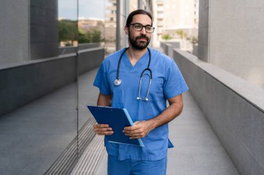 Steteskoplu mavi önlüklü erkek doktor hastane binasının dışında yürüyordu.