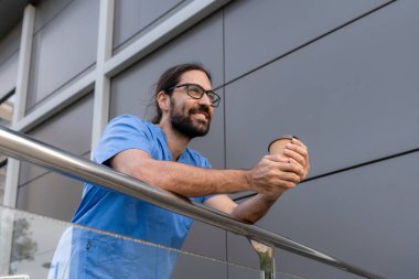 Gülümseyen erkek doktor hastane binasının dışında mola veriyor kahvenin tadını çıkarıyor ve başka tarafa bakıyor.