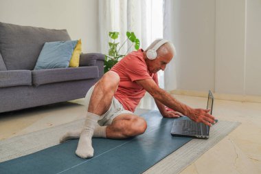 Son sınıf öğrencisi yoga minderinde oturuyor. İnternetten dersini takip ediyor ve evde kulaklık takıyor.