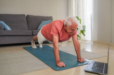 Üst düzey bir adam laptoptaki beden eğitimi dersini takip ederken yoga minderinde kalas egzersizi yapıyor.