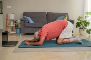 Son sınıf öğrencisi evde dizüstü bilgisayardaki beden eğitimi dersini takiben paspasın üzerinde çocuk pozu yoga yapıyor.