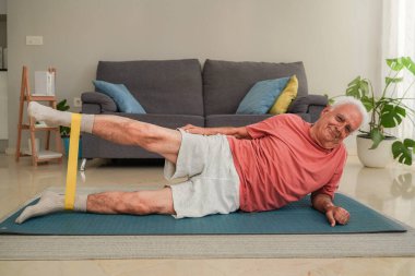 Evde yoga minderinde yatan direniş bandıyla bacak hareketliliği ve dayanıklılık eğitimi alan yaşlı bir adam.